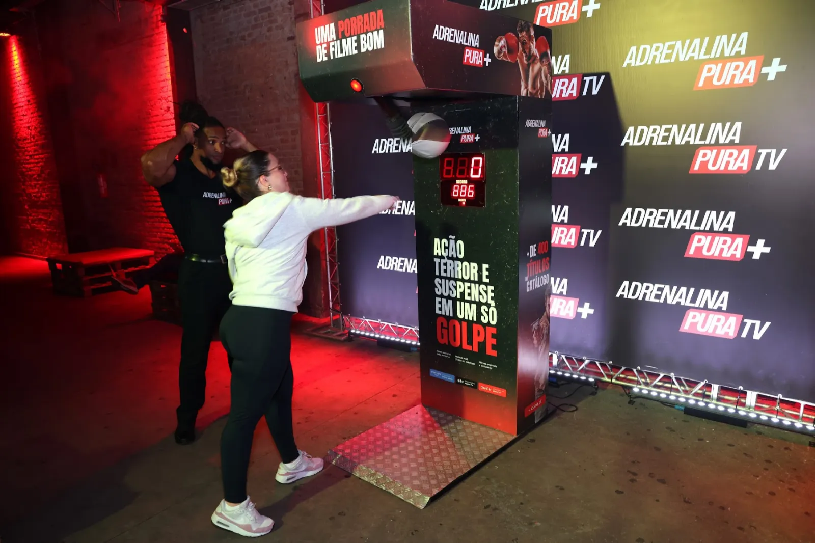 Convidada testa a máquina de soco temática do Adrenalina Pura+, em frente ao backdrop da marca.